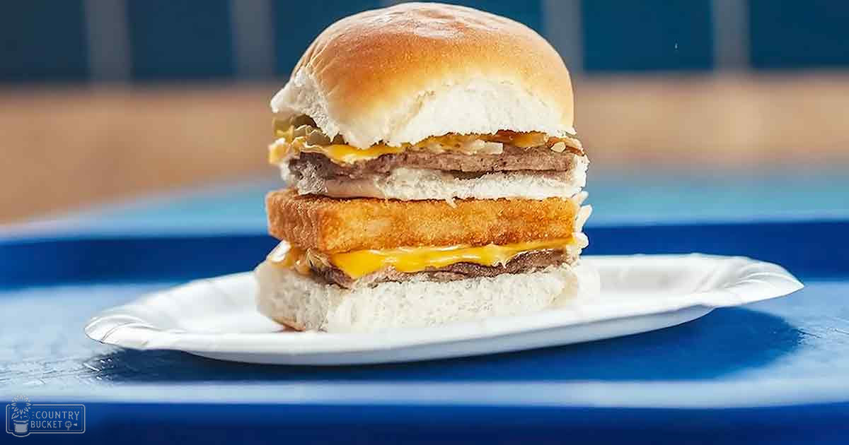 A plated burger with two patties, cheese, and a fried chicken patty or similar ingredient on a white paper plate over a blue surface indoors in a casual setting.