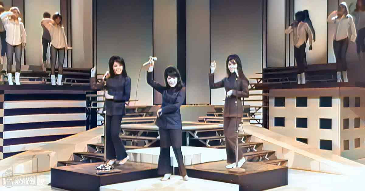 A theatrical performance set on an indoor stage with modern geometric backdrop and performers wearing similar black outfits, holding microphones. The scene is illuminated by stage lights.