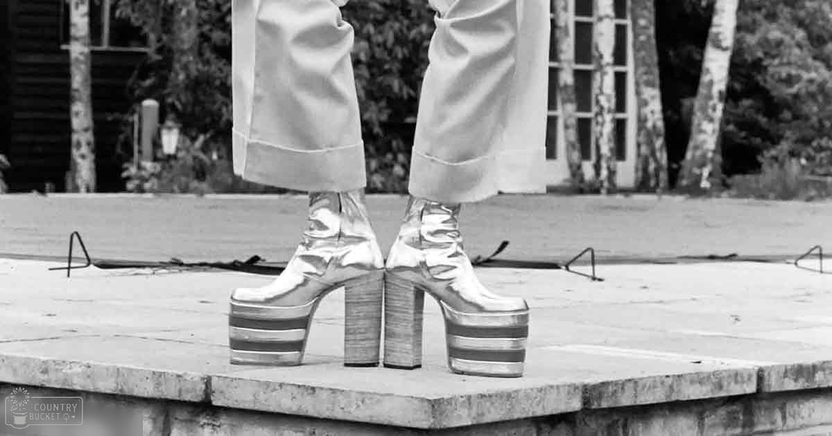 Adult female wearing silver platform boots, standing outdoors on a wooden stage with trees and buildings in the background.