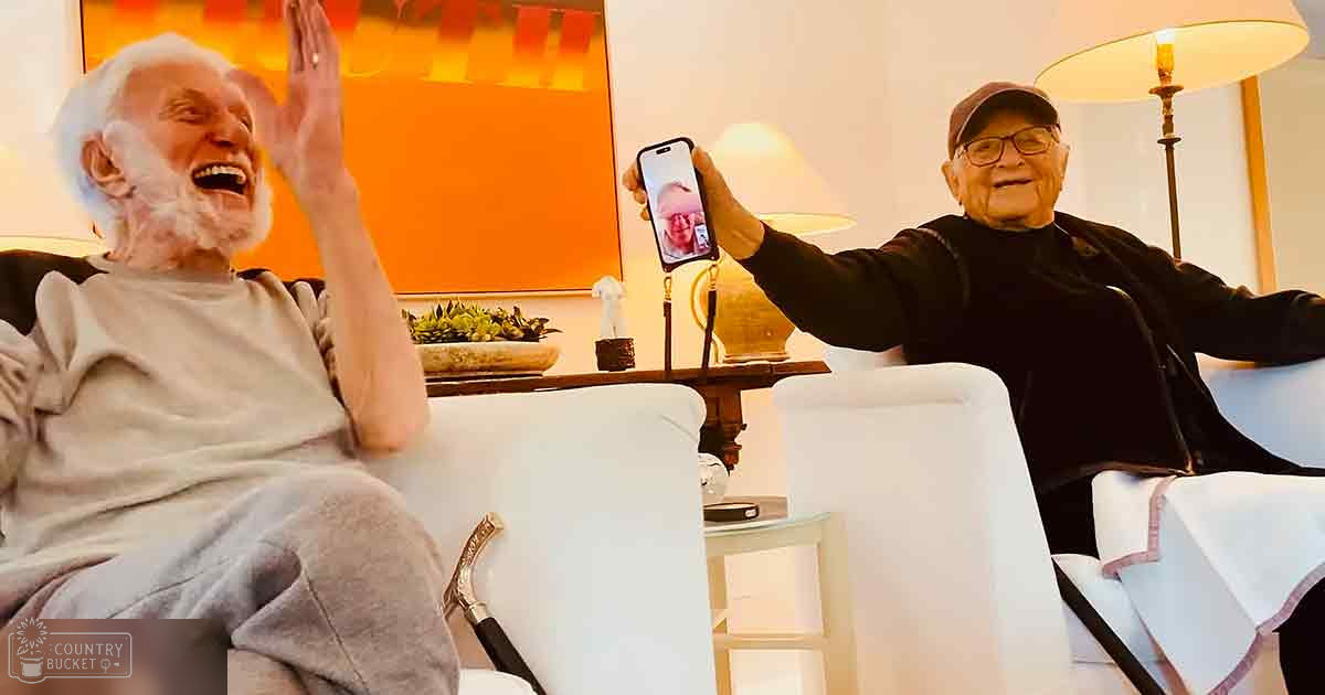 Two older adults in a cozy living room setting, one holding up a smartphone displaying another person's face.