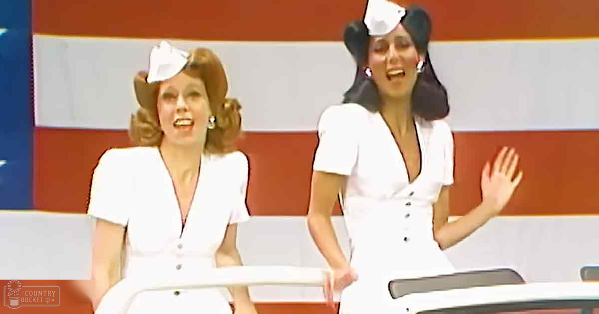 Two adult female performers wearing white dresses with V-necklines and bows, seated against a red and white striped backdrop resembling an American flag in a staged performance setting.