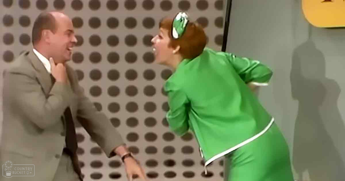 A comedic scene from a television show with an adult male in a light gray suit with a white shirt and dark tie, alongside an adult female wearing a bright green outfit with white trim and a white belt at the waist. The backdrop resembles soundproofing material commonly found in recording studios or TV sets.