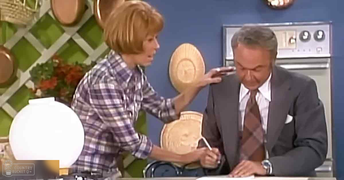 A young adult female wearing a plaid cotton or polyester blend shirt interacts with an older male in a modern kitchen-themed TV studio set, with decorative green lattice patterns and hanging copper pots. A round wooden plate is being held by the woman.