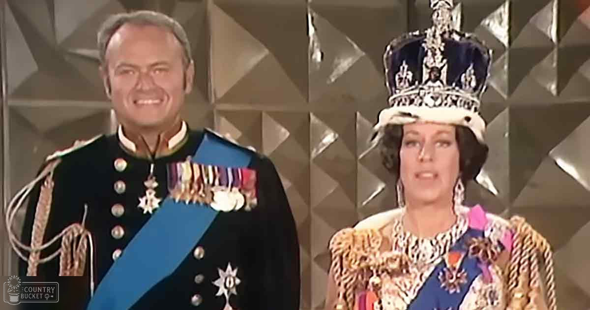 A middle-aged male in a black military-style uniform with medals and blue sash stands beside an older adult female wearing intricate gold-embroidered ceremonial gown and crown, suggesting they are part of a formal royal event. The setting is indoors with a geometric patterned backdrop.