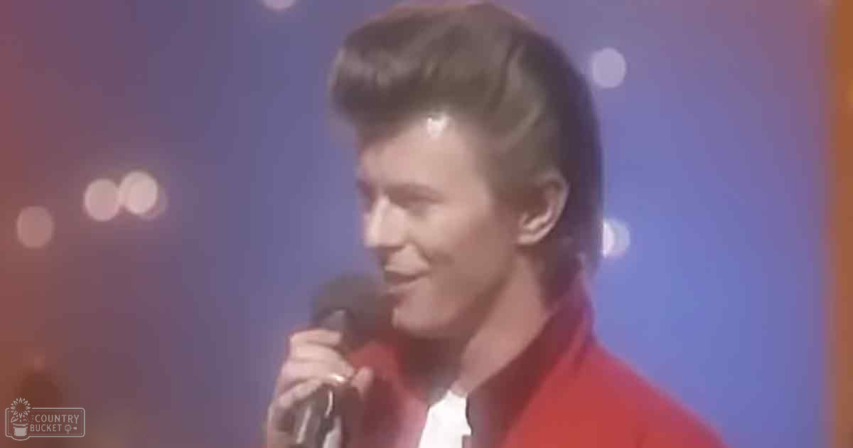 A young adult male singer wearing a red jacket with a structured collar, performing into a microphone under stage lighting at an evening concert.