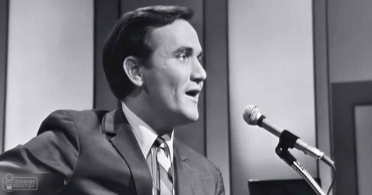 Adult male in a well-fitted suit jacket with notched lapel, collared shirt, and tie, standing indoors near microphone stand.