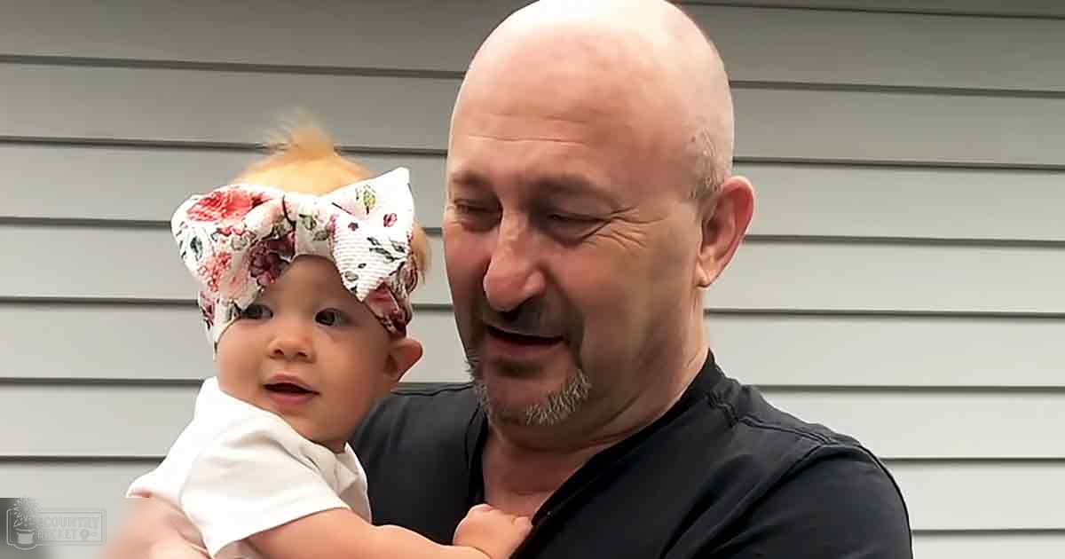 A middle-aged man holds an infant in front of a residential house, wearing a dark-colored short-sleeved shirt and the infant is dressed in light clothing with a floral-patterned bow headband. The scene suggests a late afternoon setting with soft natural lighting.