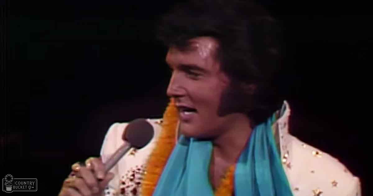 An adult male performer in a mid-20th century stage setting, wearing a white jacket adorned with gold stars and sequins, holding a microphone.