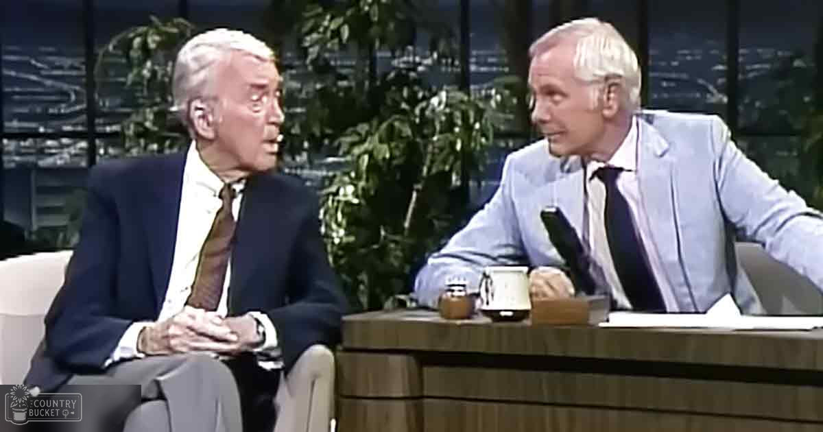 Two older adults conversing in a talk show setting with indoor plants and an out-of-focus cityscape at night.