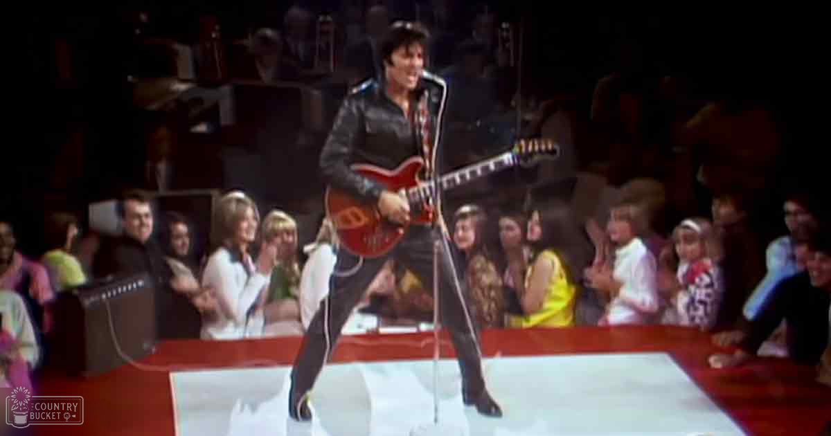 Adult male performer playing electric guitar on stage wearing black leather jacket over red shirt with dark pants and white sneakers; audience includes a toddler and children, red stage, microphone stand, amplifier, 1960s or early 1970s style setting under artificial lights.