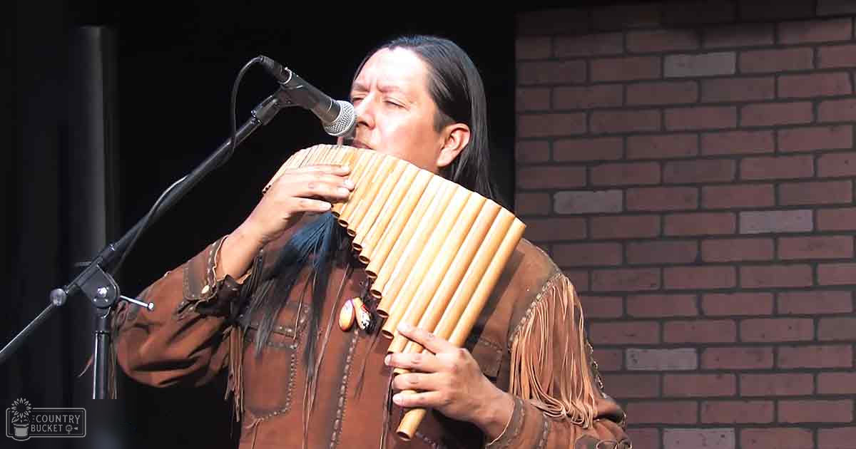 Adult male musician performing with a pan flute in an outdoor setting, wearing a brown leather jacket with fringes, under natural daylight.