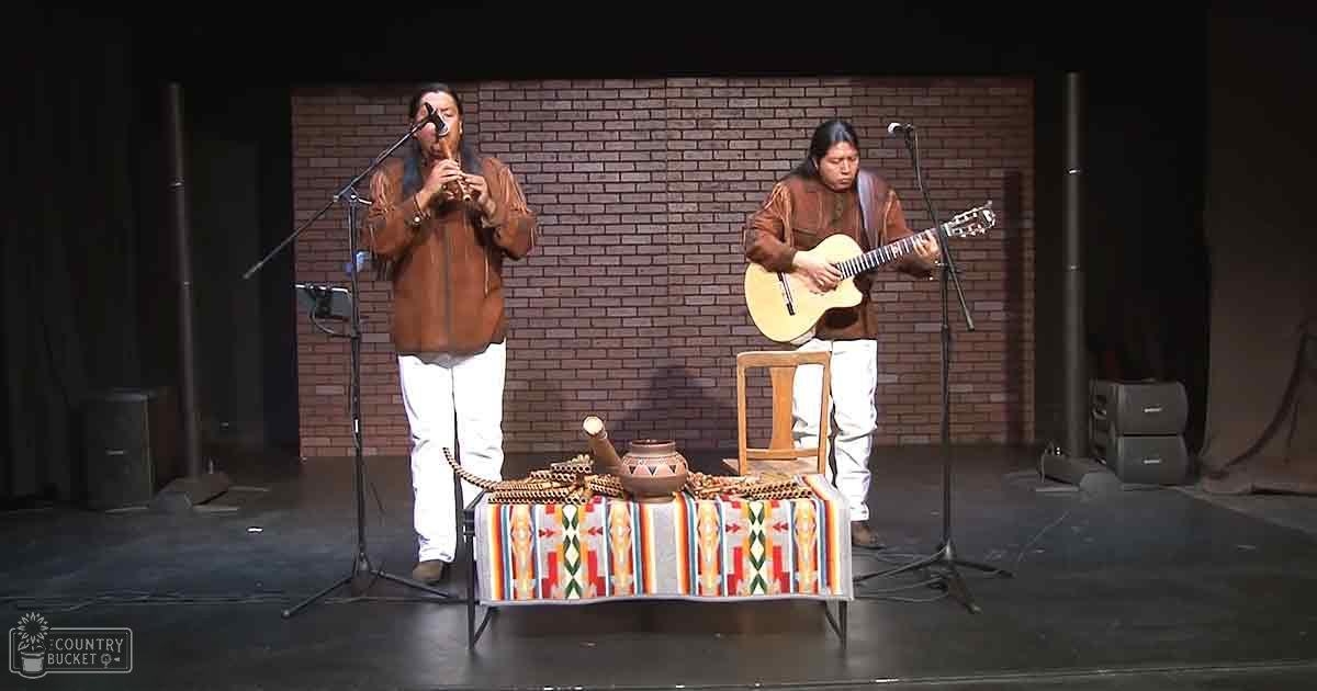 Musical performance on an indoor stage with a simple brick wall backdrop and performers wearing brown jackets and white pants.