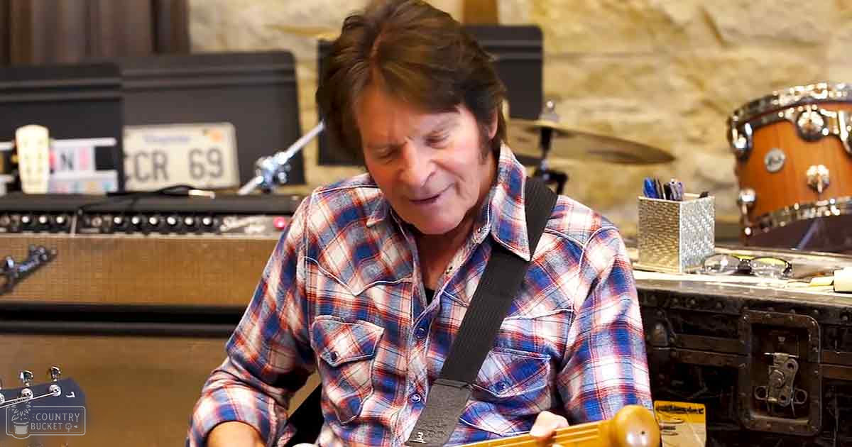 A middle-aged adult male wearing a blue, red, and white checkered shirt plays guitar in what appears to be a music studio or rehearsal space with visible musical equipment and amplifiers.