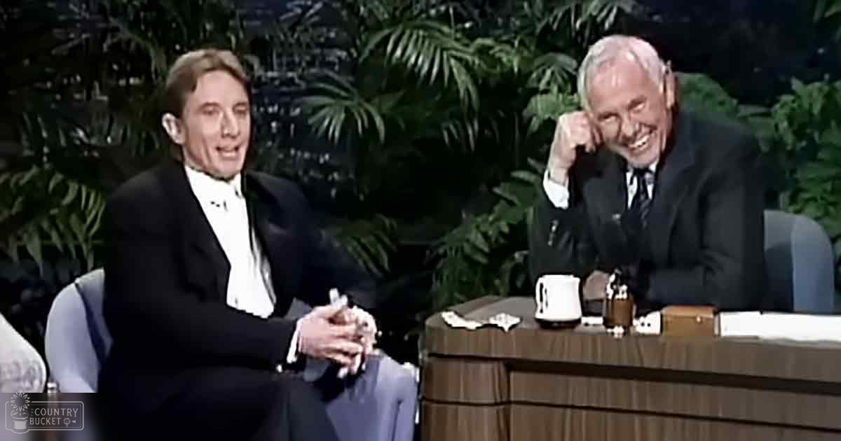 Indoor talk show set with two adult males seated at a round wooden desk, lush green foliage and artificial lighting in the background.