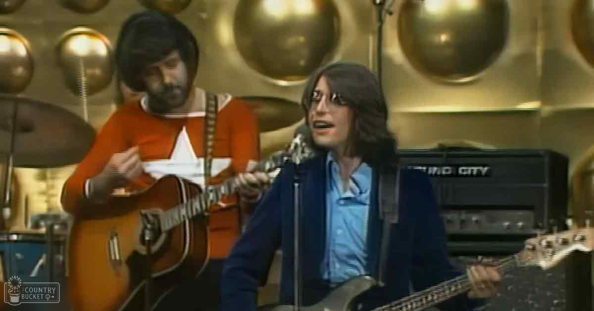 A young adult male musician in a music studio, wearing a blue shirt with dark blue cardigan and glasses, playing an electric guitar. Another adult male musician plays an acoustic guitar in the background.