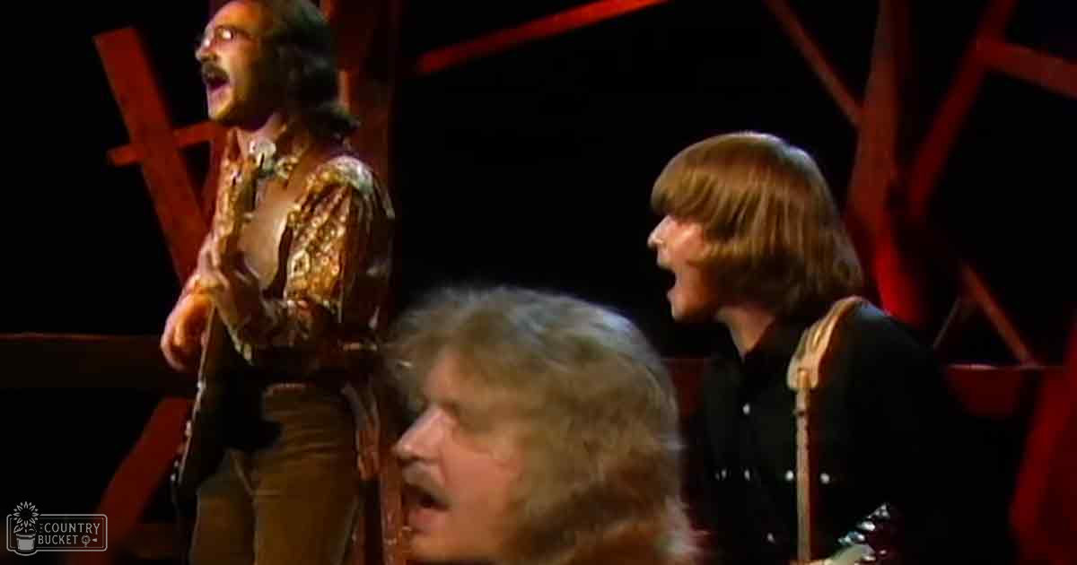 A musical performance scene indoors on stage at night, with three adult males in vintage and solid-colored shirts under dim red-lit conditions.