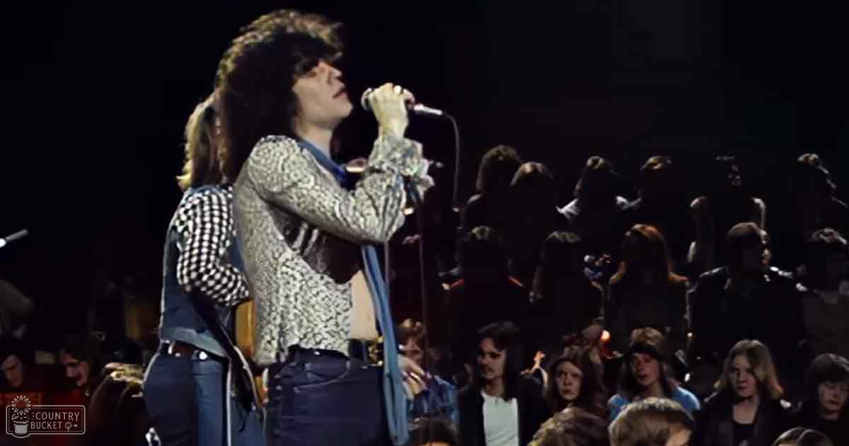 A young adult male singer wearing a patterned long-sleeved shirt performs into a microphone at a concert with various audience members in casual attire, under stage lighting.