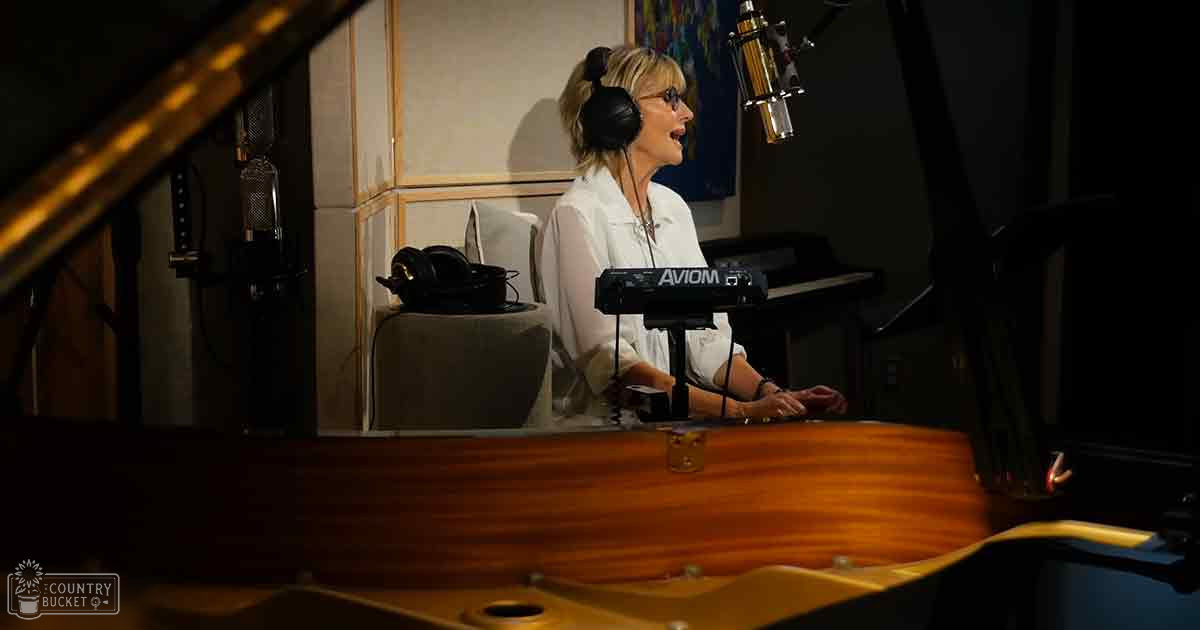 A middle-aged adult female in a recording studio, wearing a white blouse with long sleeves and black headphones around her neck.