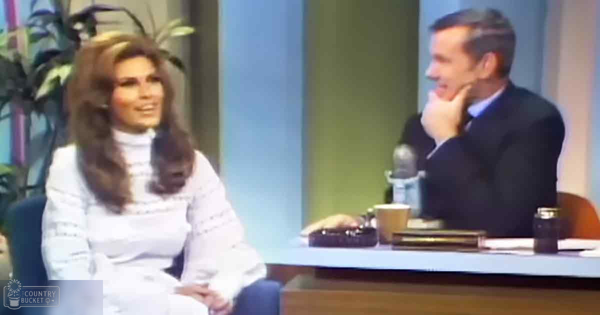An adult female seated during an interview, dressed in a vintage white turtleneck sweater and skirt from the 1960s or early 1970s, with an older male interviewer in a dark suit jacket and light blue collared shirt, seated across her at a desk in a professional studio setup typical of daytime television productions.