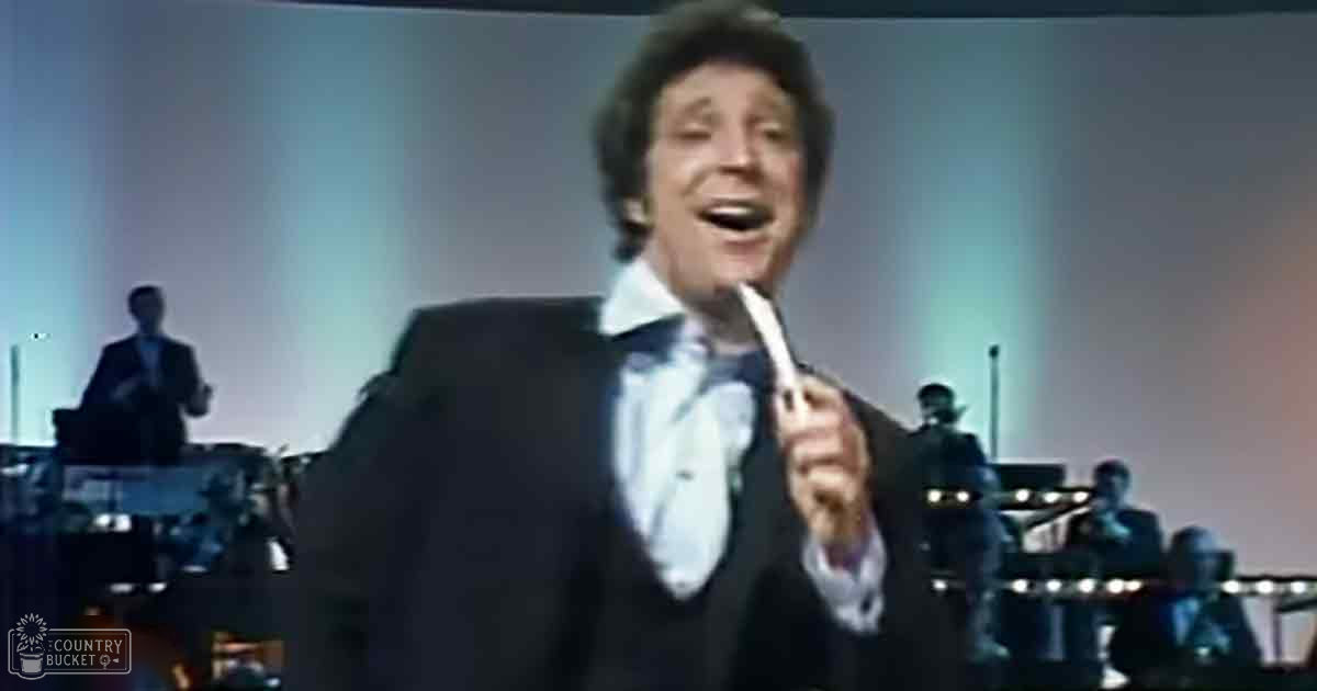 A middle-aged male performer in a dark suit jacket with white shirt and bow tie singing into a microphone on stage during what appears to be a live concert performance. Background musicians, one playing drums and the other keyboard, are also present.