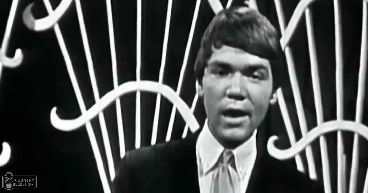 A young adult male in a well-fitted dark suit jacket, crisp white shirt, and tie stands indoors against ornamental wrought iron; mid-20th-century style.
