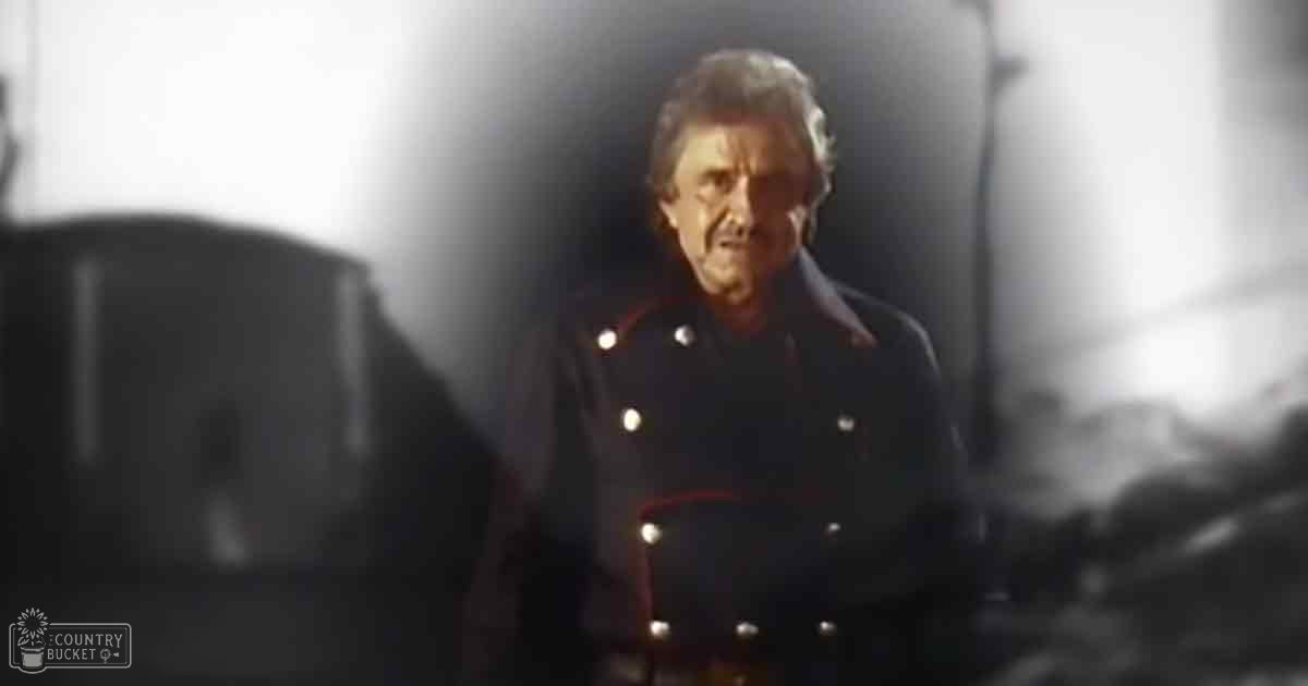 A middle-aged man in a structured, dark-colored military-inspired jacket stands indoors holding a guitar; text reads 'Madly Odd' in the corner.