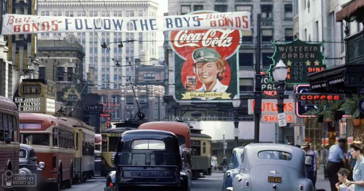 A bustling mid-20th century city street scene with vintage cars, a Studebaker sedan, trolleybus and regular bus, and advertisements for bonds, Coca-Cola, Loews theater marquee, and Butlers Shoes. The architecture suggests San Francisco.