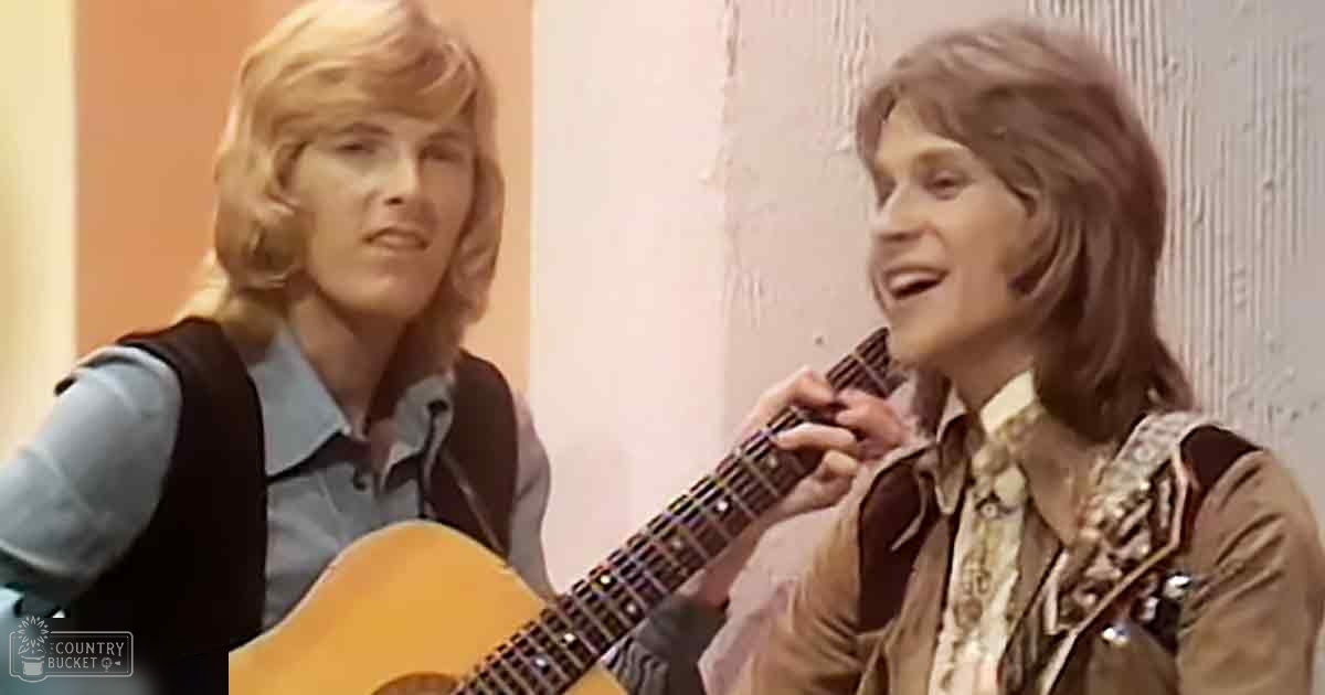 Two young adult males in a casual indoor setting, wearing light blue shirts with brown vests and patterned white shirts layered under jackets, playing guitar against plain wall background with discoloration.
