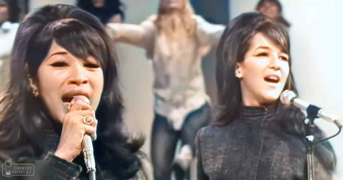 A musical performance by two young adult female singers dressed in mid-20th century-style jackets, held outdoors with a crowd of people in the background and late afternoon sunlight casting shadows.