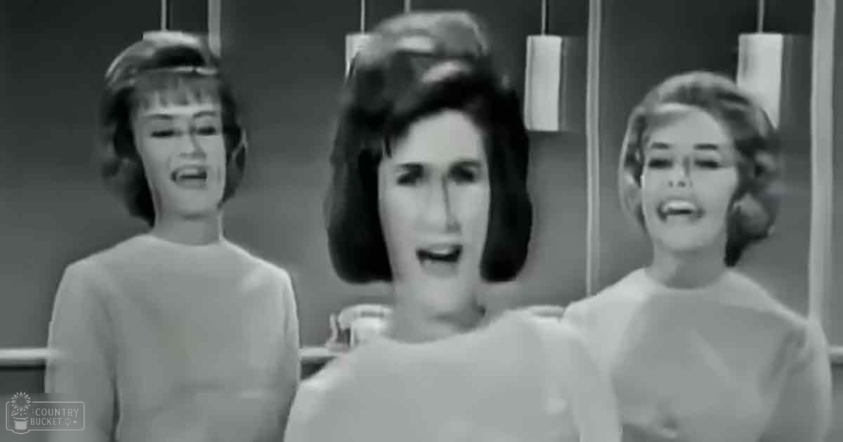 Three women singing in an indoor studio setting with a backdrop of vertical bars.