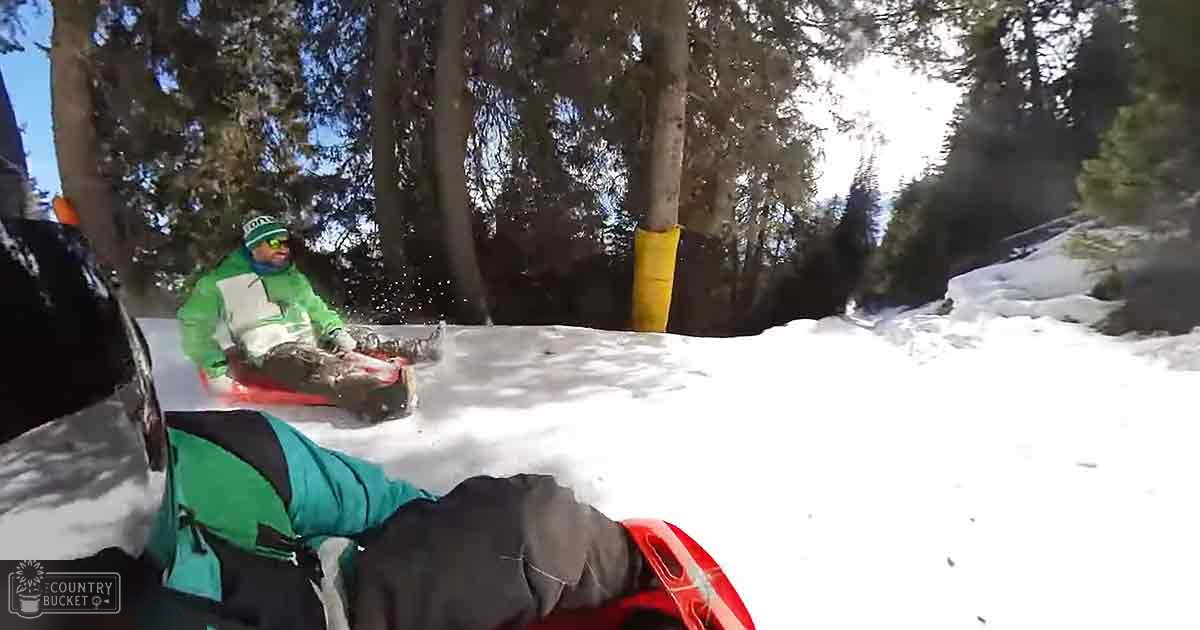 A child in a bright green jacket and matching hat sleds down a snowy hill with an adult supervising, surrounded by tall evergreen trees under bright daylight.