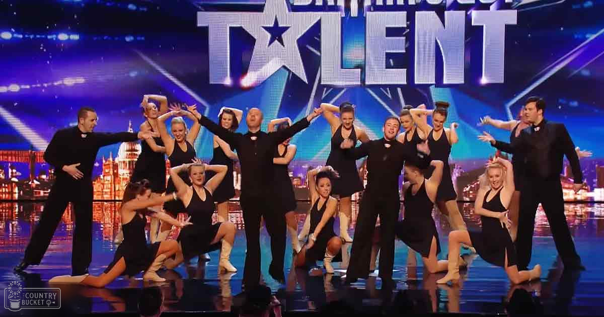 A dance troupe performing at 'Britain's Got Talent' with male dancers in black suits and female dancers in form-fitting black dresses on a stage with an illuminated cityscape background.