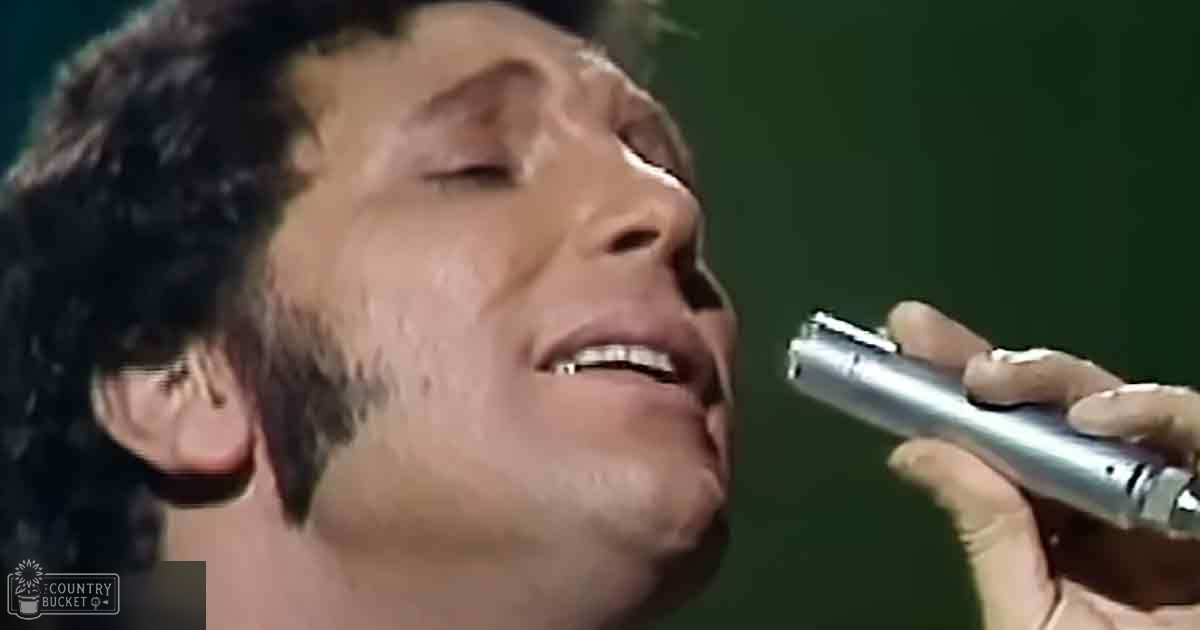 An adult male singer in a mid-20th-century-style dark-colored garment with long sleeves, singing into a silver microphone at an evening concert venue.