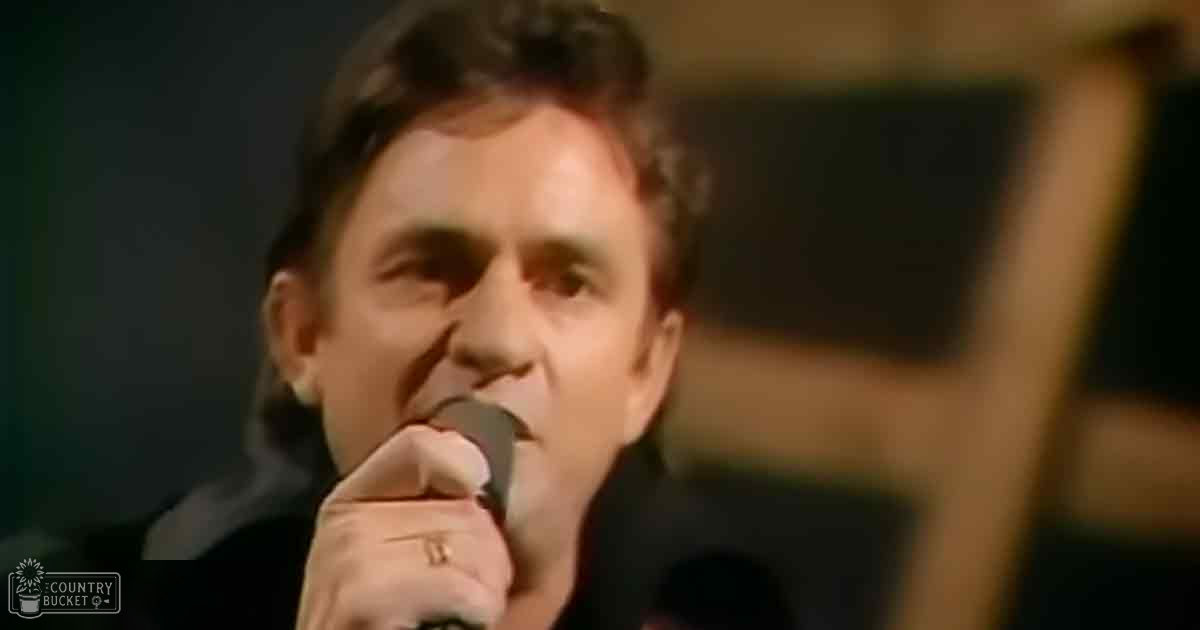 A middle-aged adult male singer wearing a dark-colored collared shirt or jacket stands on stage under dim lighting, performing into a microphone.