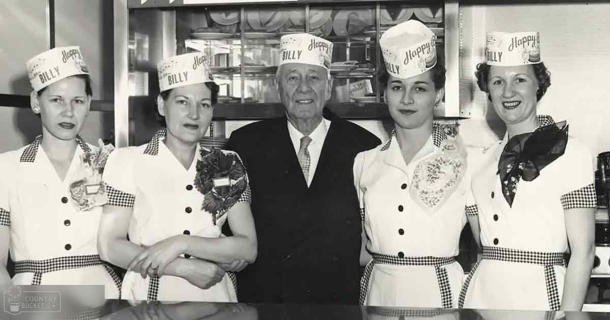 An older adult male in a dark suit and patterned tie, surrounded by individuals wearing white uniforms with black-and-white checkered trim and floral brooches, suggesting a mid-20th century professional setting.