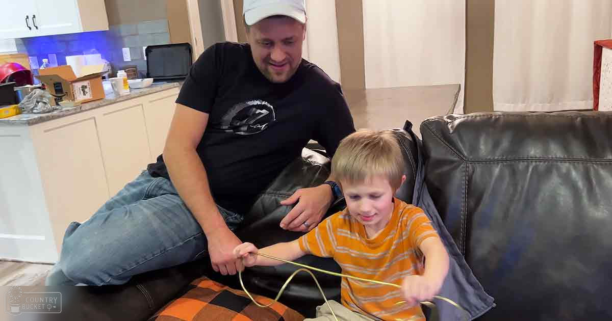 A middle-aged male wearing a black t-shirt with a graphic design, blue jeans, and a white cap stands in a living room with a child around 5-7 years old in an orange striped shirt and beige pants. The setting includes a black leather couch with a plaid pillow and kitchen cabinets, countertops, and various household items.