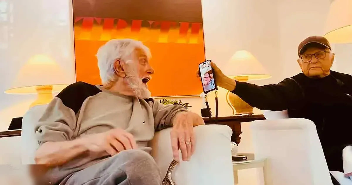 An older adult male in a living room setting, wearing a gray hoodie and holding up a smartphone.