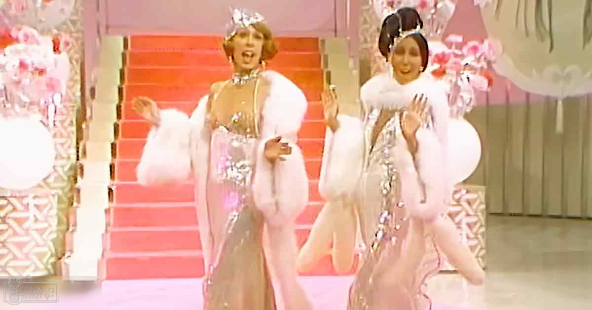An adult female singer in an elegant white gown with pink and silver accents, adorned with a matching ornate headpiece, on stage with red carpeting and decorative floral arrangements.