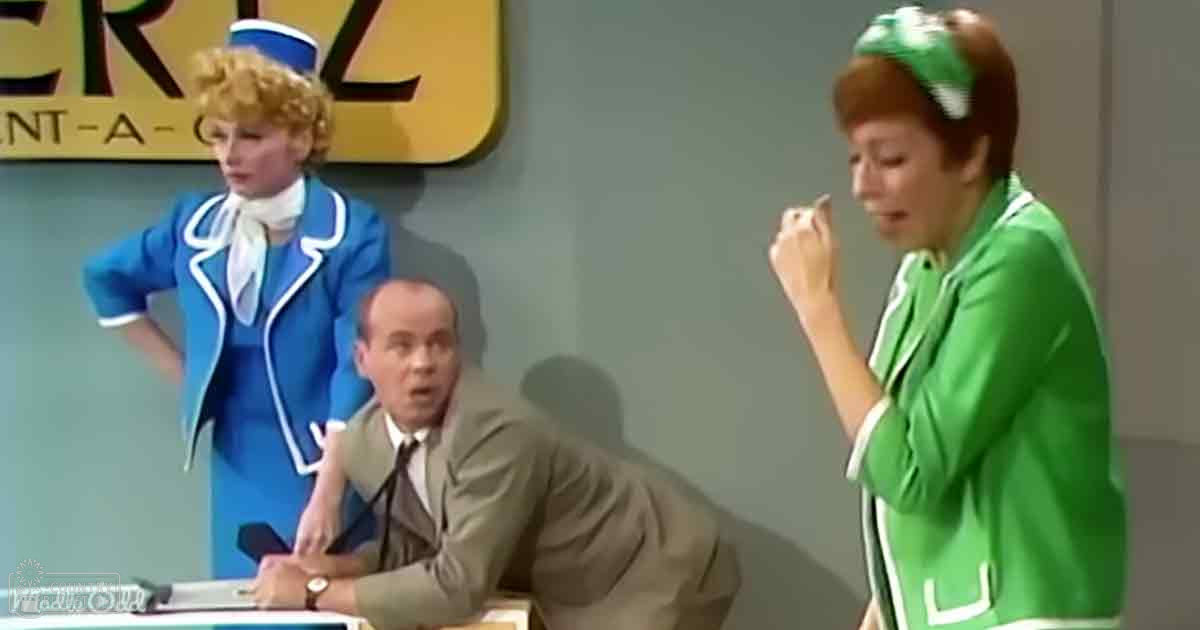 Adult male in light brown suit and white shirt seated at desk; adult females in blue and green uniforms stand nearby, one wearing a white scarf and small hat, the other with a green headband; 'Hertz Rent-A-Car' sign visible on wall behind woman in blue; indoor office setting with neutral walls and bright lighting.