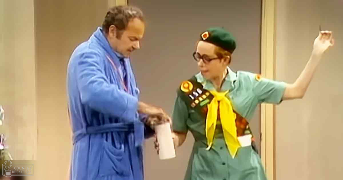 A middle-aged male in a blue bathrobe interacts with a young adult female wearing a green uniform and beret, possibly during a comedy sketch indoors.