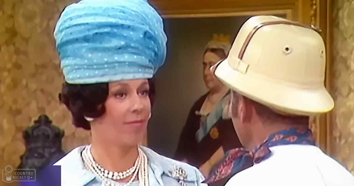 Adult female wearing a light blue jacket with a brooch and pearl necklace, seated indoors near an adult male in a beige pith helmet and white shirt.