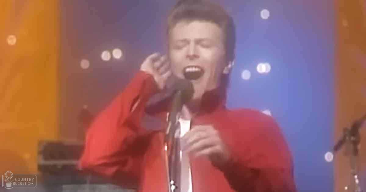 A young adult male singer performs on stage at a concert, wearing a red jacket and white shirt in an indoor setting with circular spotlights.