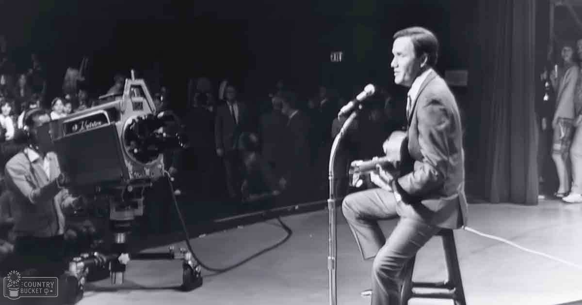 A middle-aged adult male performer wearing a mid-20th-century style suit jacket and trousers sits on stage with a guitar, singing or speaking into a microphone at what appears to be a television studio set.