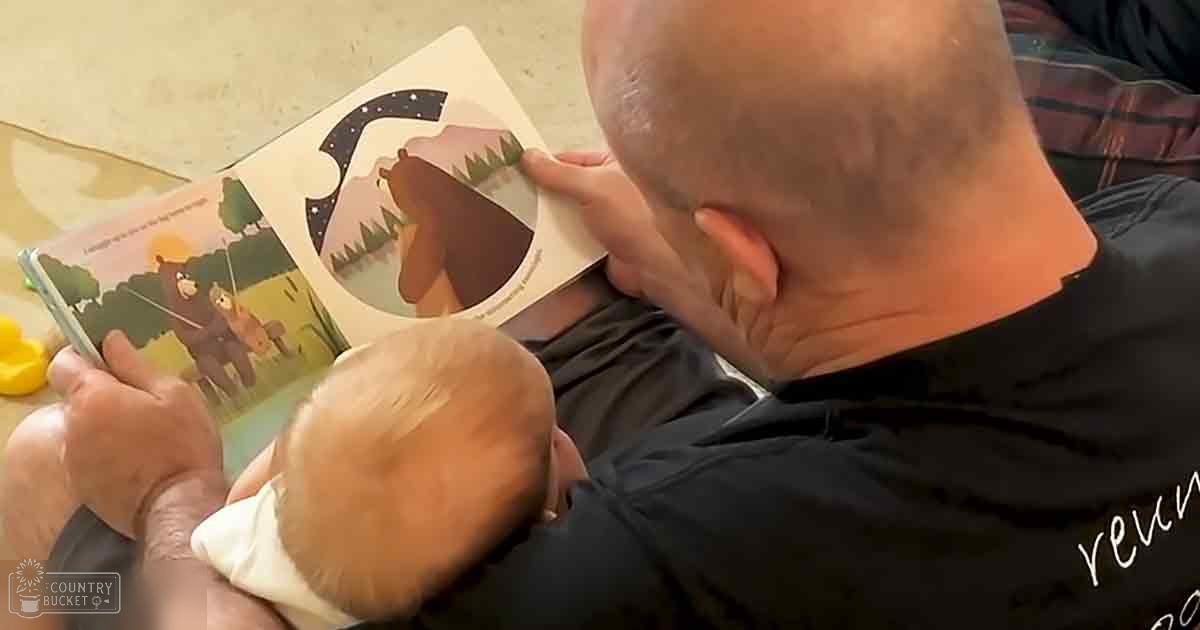 A middle-aged man reads to an infant in a home setting, both wearing casual clothing. The focus is on the open children's book with illustrations of bears and trees.