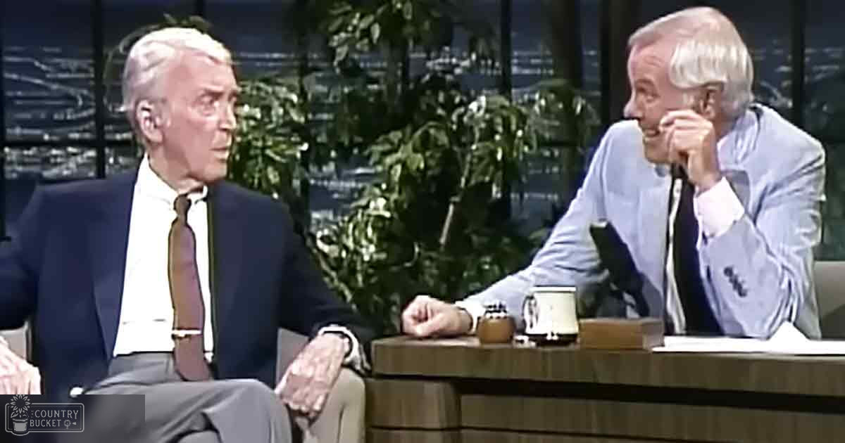 Two older adult males in a talk show setting, seated at a desk with city lights visible through large windows at night.