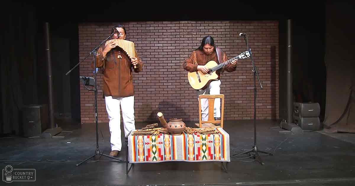 An indoor musical performance with two adult performers on stage in front of a brick wall backdrop under artificial lighting.