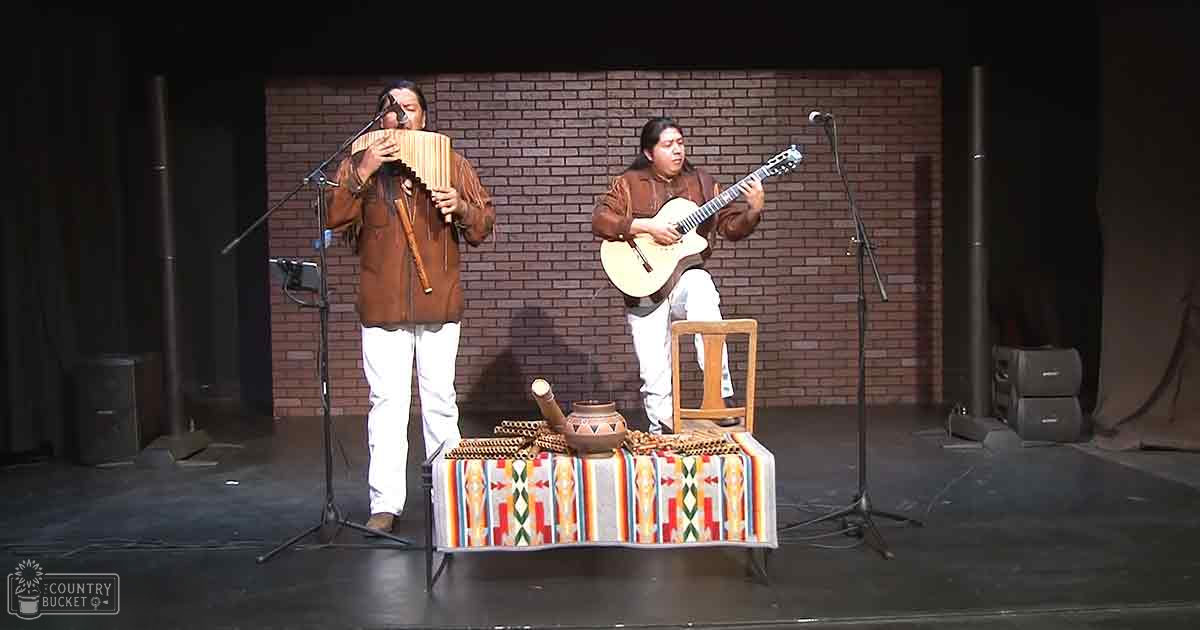 Musical performance in an indoor setting with two adult male performers wearing brown leather jackets and white pants on a stage with brick wall backdrop and patterned tablecloth.