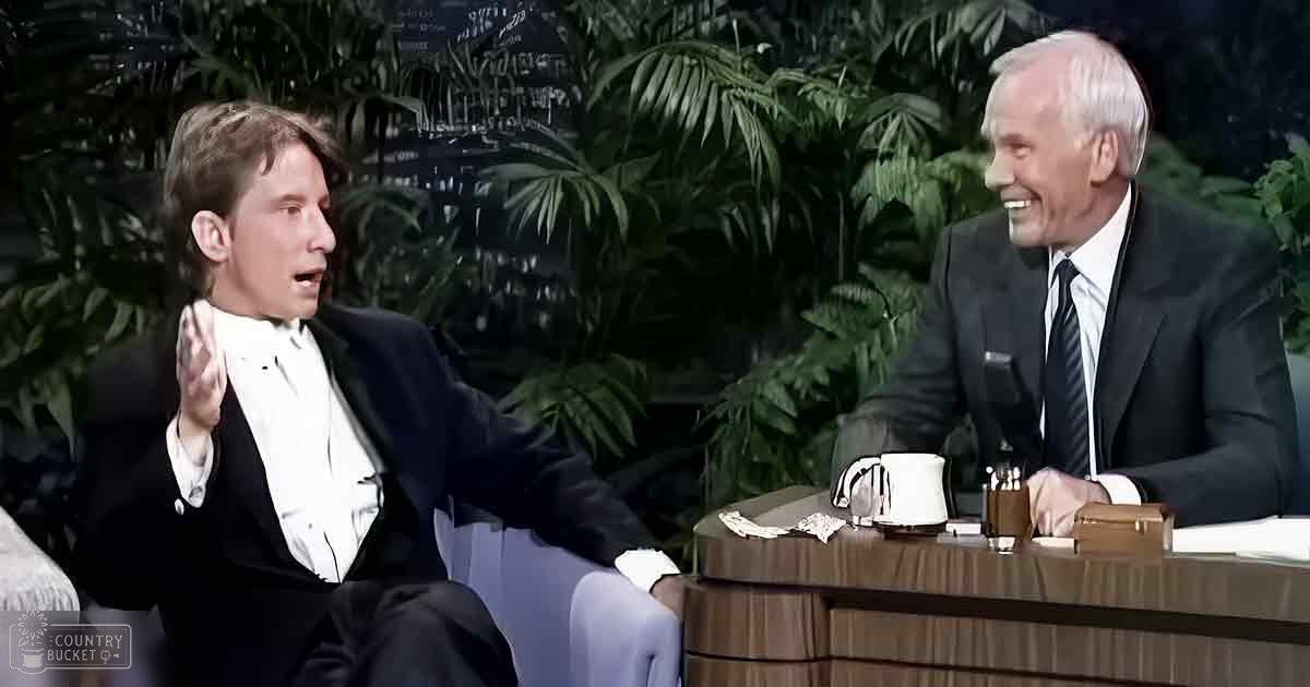 Two adults in a talk show setting, one older male in a well-tailored dark suit and another adult male in a formal suit, surrounded by indoor plants and studio furniture with evening lighting.