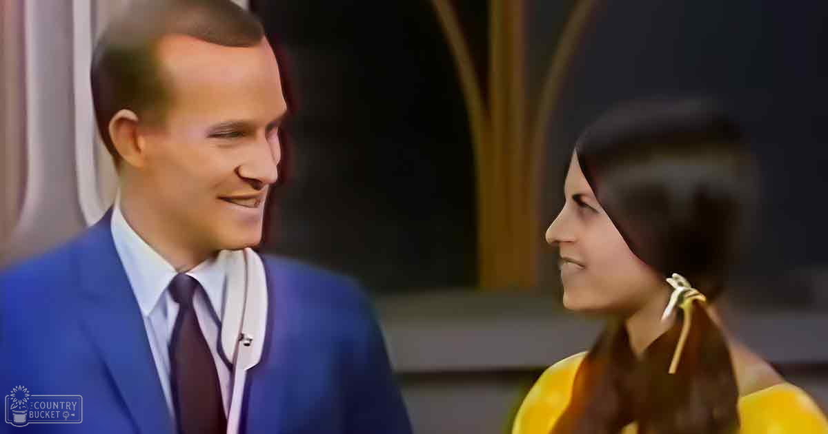 Adult male in a blue suit jacket and white shirt with dark tie stands indoors alongside a young adult female wearing a yellow dress with pigtails tied with white ribbons, the setting appears to be an interior space possibly formal or semi-formal.