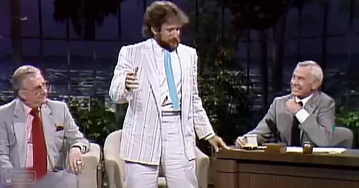 Adult male in a white striped suit jacket with a microphone, seated men in formal attire, indoor studio setting with beige armchairs and greenery outside.